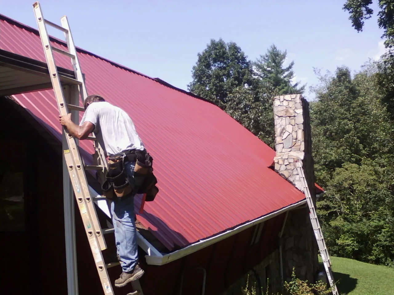 Roofer on a ladder beside a red metal roof and stone chimney — Soffit & Fascia Repair by Roofing Brothers in North Liberty